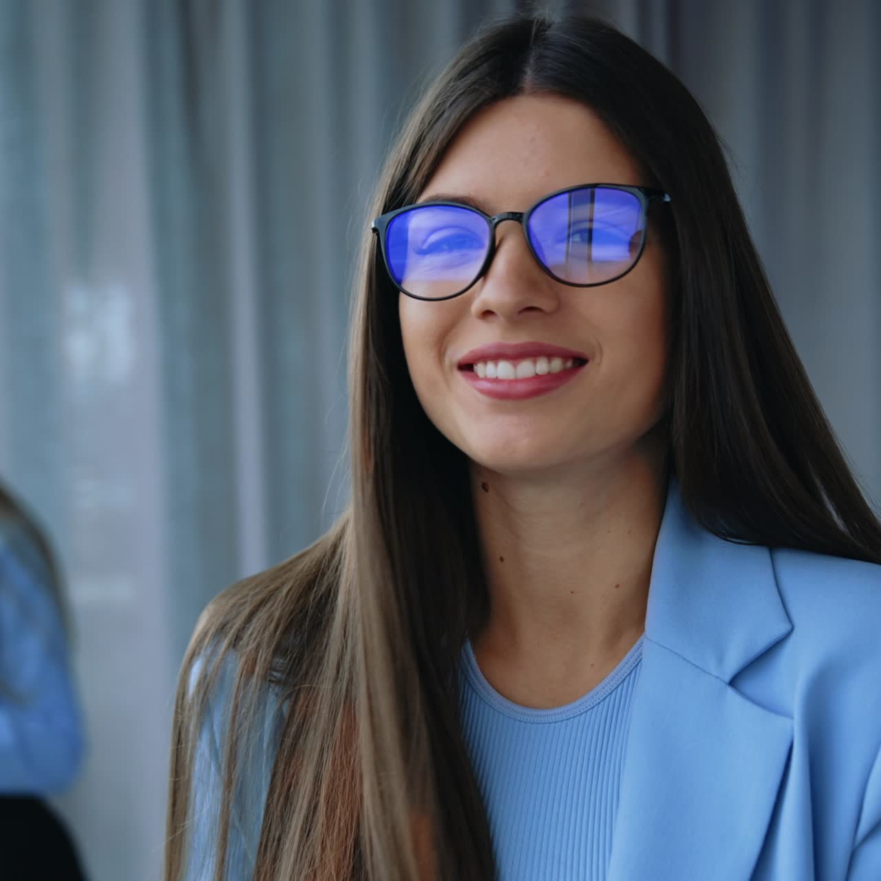 Good-looking young long-haired lady wearing blue jacket stands at the desk in office. Business woman puts on glasses smiling to camera. Another lady holding phone standing at window at backdrop in blur