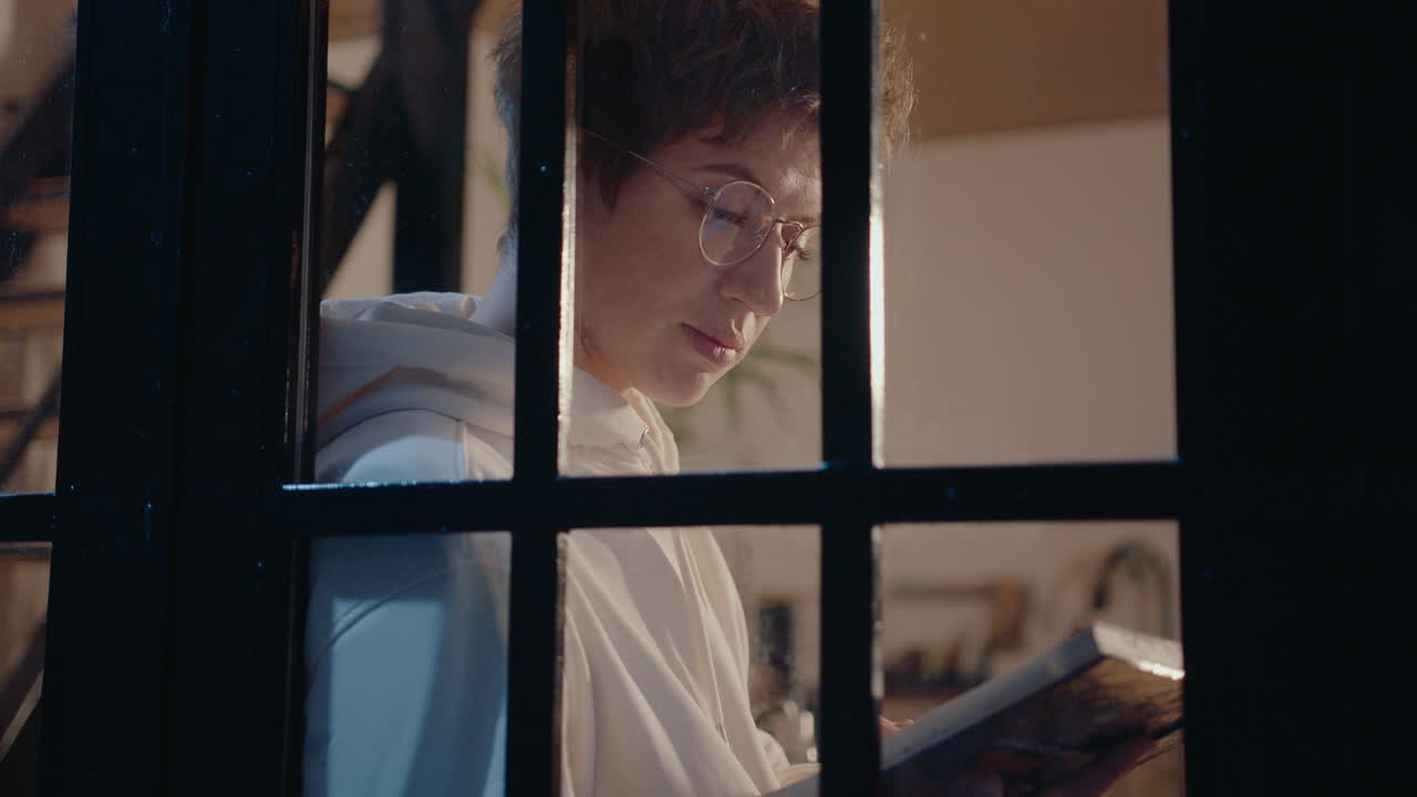 Young Woman Reading Book by Window in the Evening