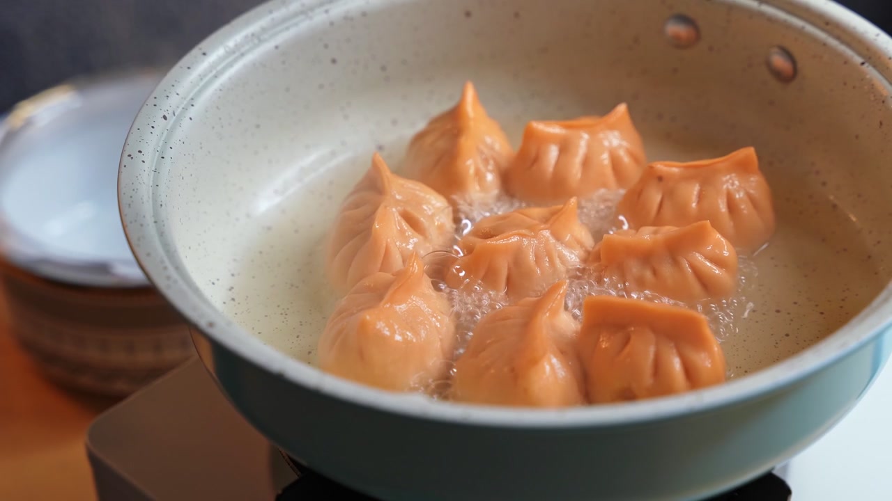 Close-up saat membuka wajan antilengket biru muda berisi pangsit oranye mengepul. Makanan panas itu dimasak dalam cairan mendidih di atas kompor portabel putih selama persiapan makan siang di dalam ruangan