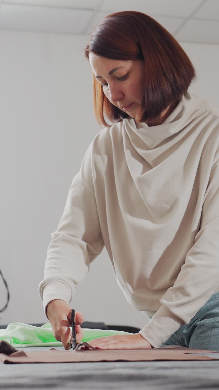 Costurera caucásica cortando tela marrón, corte meticuloso de patrones sobre una mesa de trabajo con unas tijeras grandes, piezas de patrón extendidas, luz de estudio neutra, expresión concentrada, ambiente de moda sostenible y de proximidad