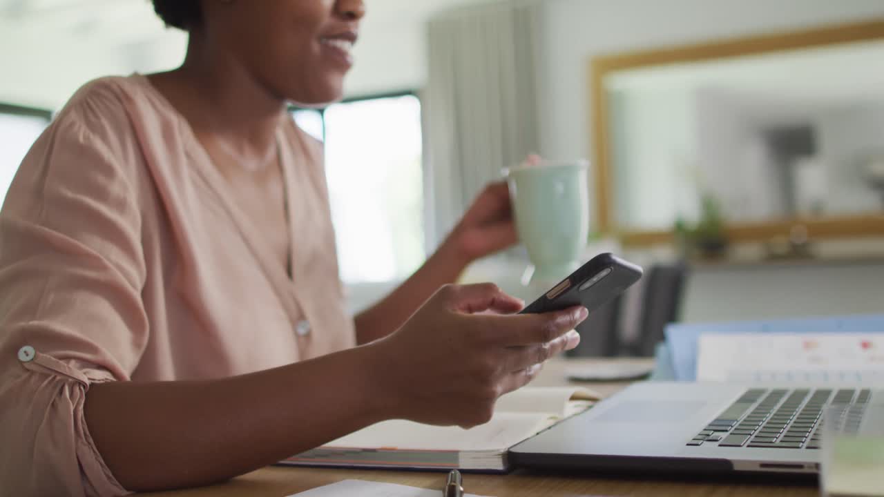 feliz mujer afroamericana sentada en la mesa usando un teléfono inteligente y bebiendo café