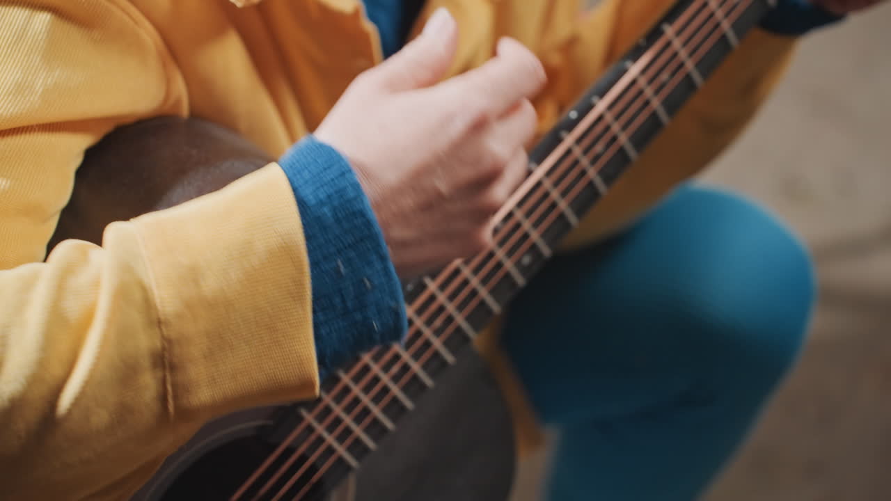 Primo piano di mani che strimpellano una chitarra acustica, atmosfera da campeggio. Giacca gialla e leggings blu incorniciano una ripresa ravvicinata di tastiera e corde. Il movimento delle dita e il polsino testurizzato del maglione creano un'intimità tattile