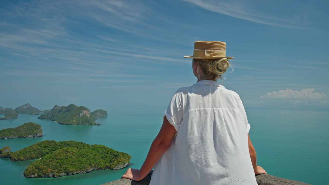 Middelaldrende kvinne med stråhatt, stående ved et naturskjønt utsiktspunkt, ser ut over turkis sjø og tropiske øyer i Ang Thong marinepark i Thailand.