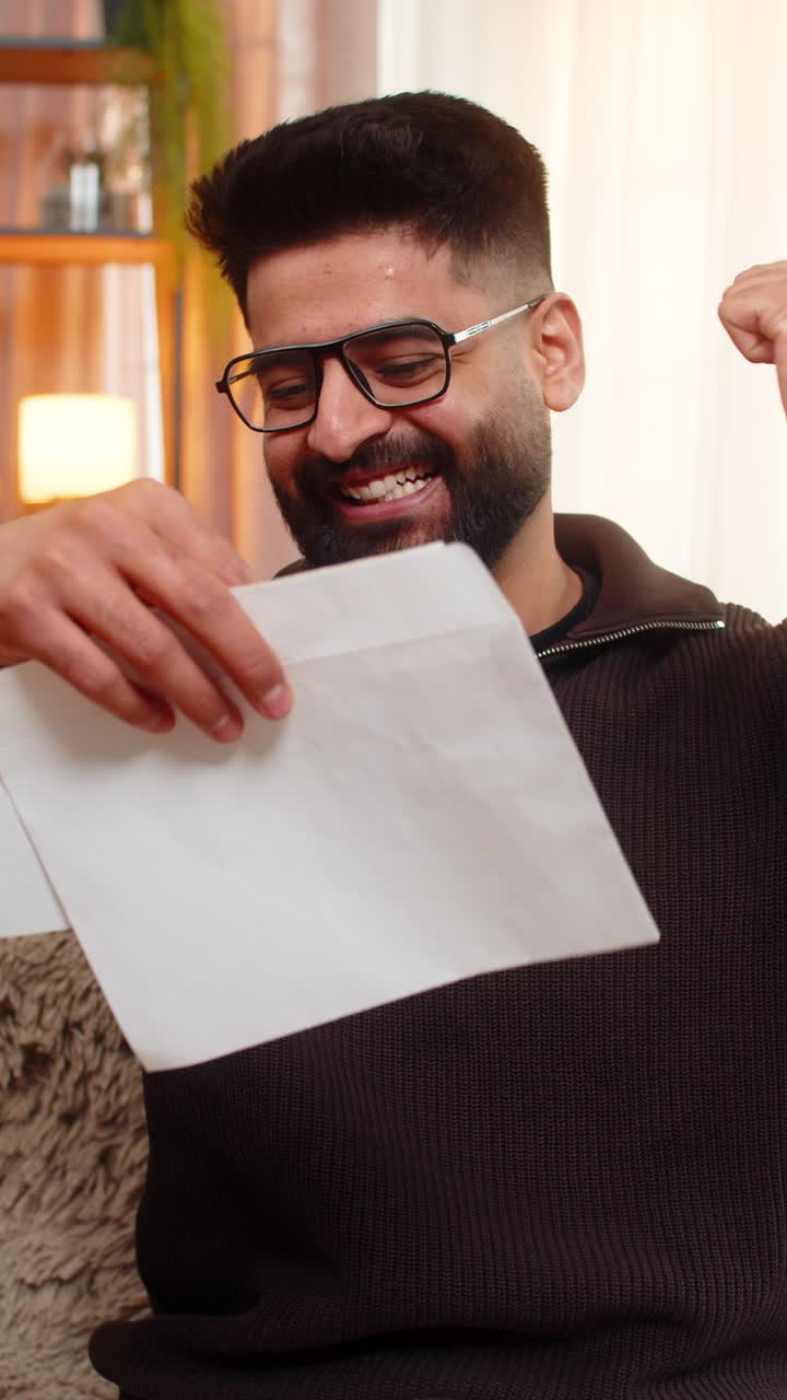 Indian man on sofa opens envelope and celebrates good news after reading positive letter at home