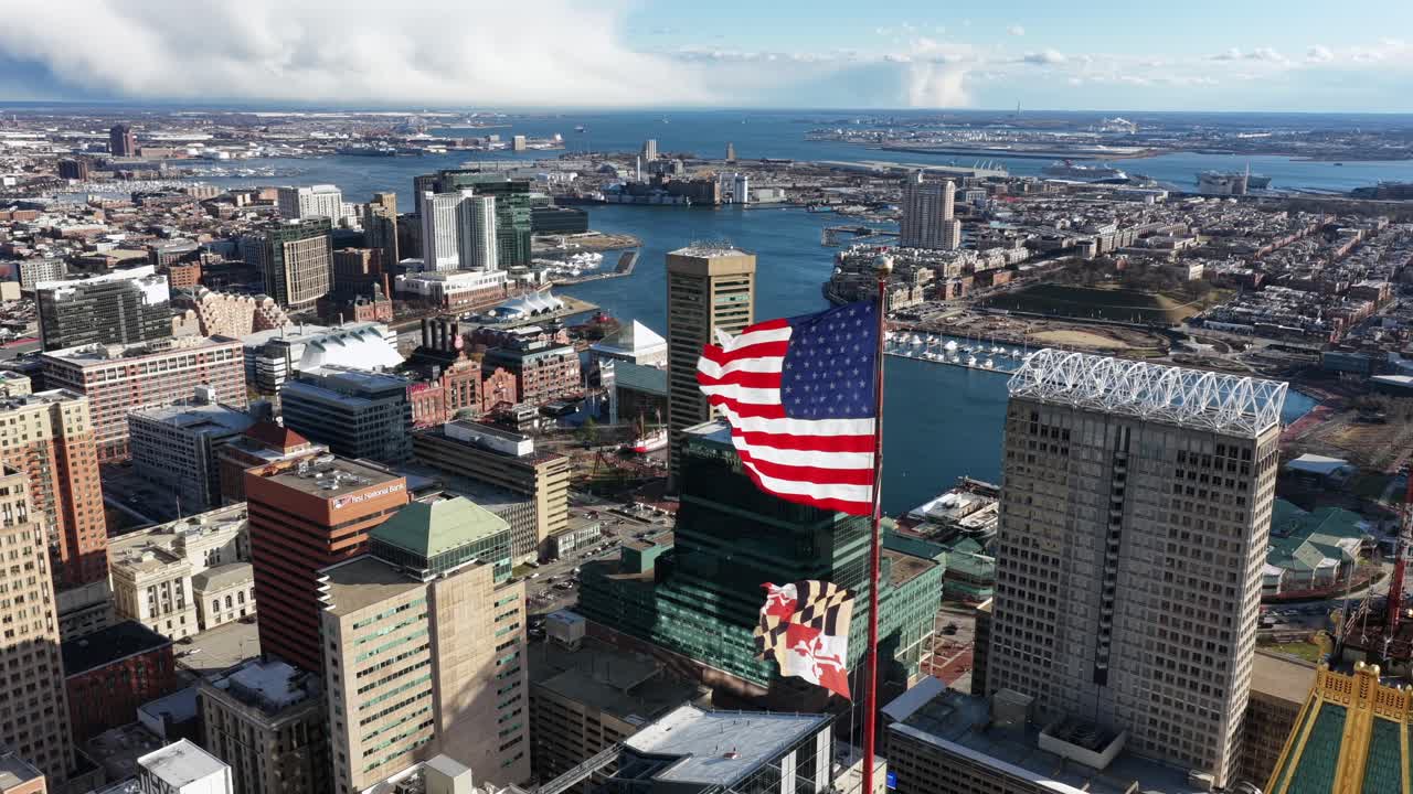 Vista aerea del centro di Baltimora con l'Inner Harbor, lo skyline dei grattacieli, il lungomare e una grande bandiera americana che sventola sopra la città in una limpida giornata di sole. Città patriottica negli USA