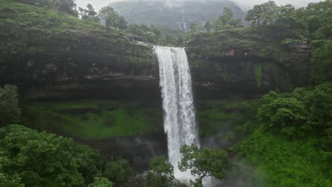 a stunning drone view of vasudhara falls in bhandardara, maharashtra india, surrounded by lush green forests and towering mountains. mystical monsoon clouds creates a serene and breathtaking ambiance