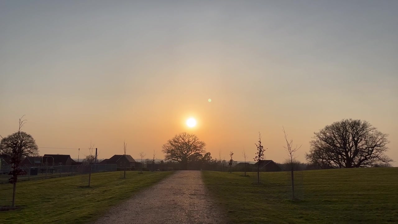 Matahari terbenam keemasan yang hangat bersinar di atas jalan kerikil melalui Knowle Park, dengan padang rumput, pohon-pohon muda, dan pohon ek dewasa yang membentuk siluet di cakrawala di Fair Oak, Hampshire, Inggris.