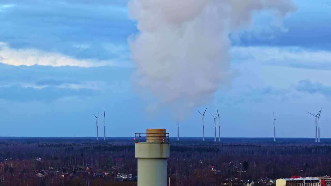 Vista aerea da drone con teleobiettivo del fumaiolo di una centrale elettrica che emette vapore bianco sui tetti dei sobborghi, con turbine eoliche all'orizzonte sotto un cielo nuvoloso e suggestivo