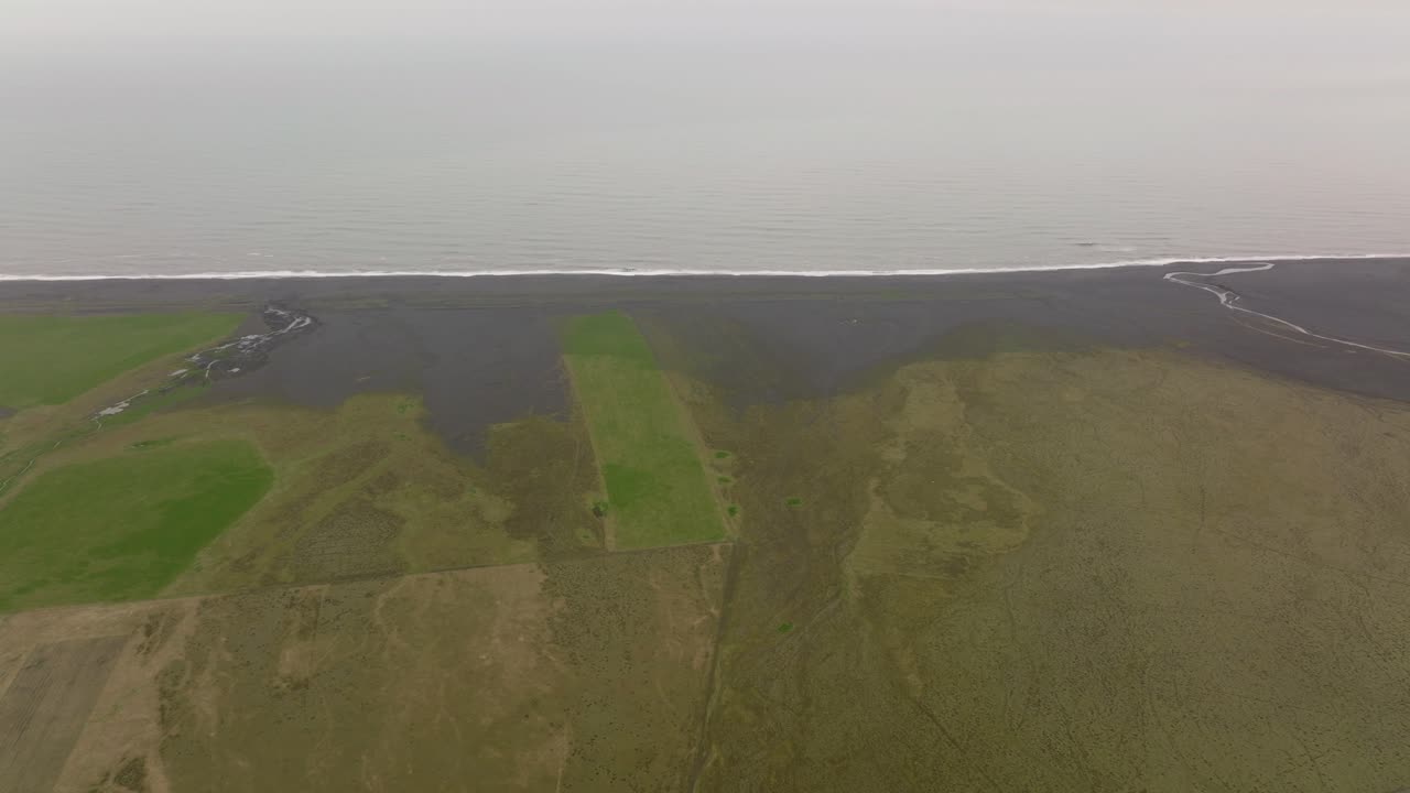 Aerial shot of coastal farmlands leading to a black sand beach near Petursey, Iceland, showcasing the contrast between lush green fields and the dark volcanic shoreline.