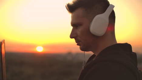 a man in headphones on the roof relaxes working remotely enjoying life despite a handsome kind of sipping beer and types on the keyboard. trade on the stock exchange using a laptop and enjoying the beautiful view