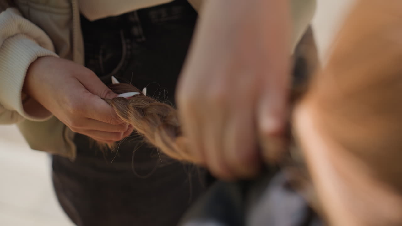 Trecce rosse vibranti intrecciate lentamente, dita illuminate dal sole che intrecciano ciocche di capelli luminose con cura, dettaglio di dita che intrecciano delicatamente capelli dai colori vivaci con luce naturale che accentua il movimento