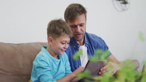hombre caucásico y su hijo sentados en un sofá en casa usando una tableta digital y sonriendo