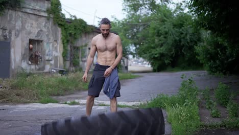 Shirtless-man-pushes-raises-large-wheel-from-tractor-outdoors