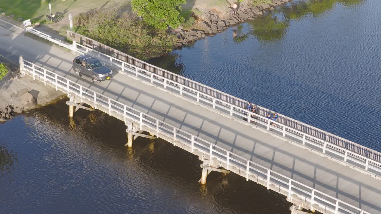 Aerial footage captures a car crossing a bridge over calm water, with clear skies and natural surroundings