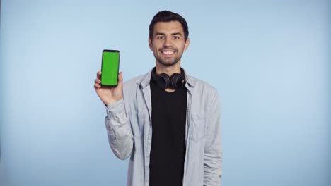 hombre guapo y curioso con camiseta azul y auriculares en el cuello que presenta un teléfono inteligente y muestra un teléfono inteligente de pantalla verde a la cámara, aislado en un fondo azul
