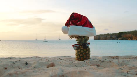 Playa-En-Curacao---Piña-Con-Sombrero-De-Navidad-Y-Gafas-De-Sol-En-La-Orilla-Arenosa-Durante-La-Puesta-De-Sol---Toma-De-Primer-Plano