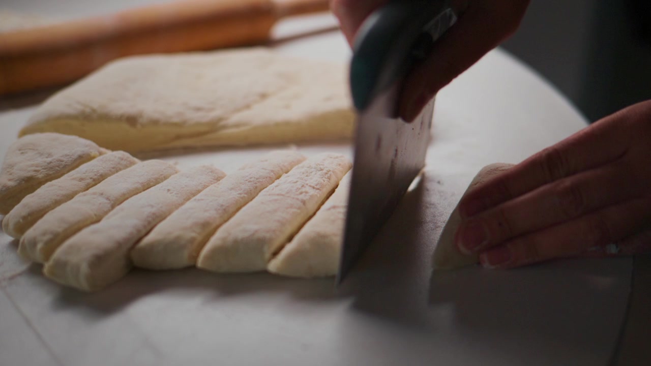 Hands slice dough into thick sticks on white surface. Rolling pin rests nearby, ready for use. Process shows careful, deliberate cutting motion. Scene evokes calm, focused cooking preparation