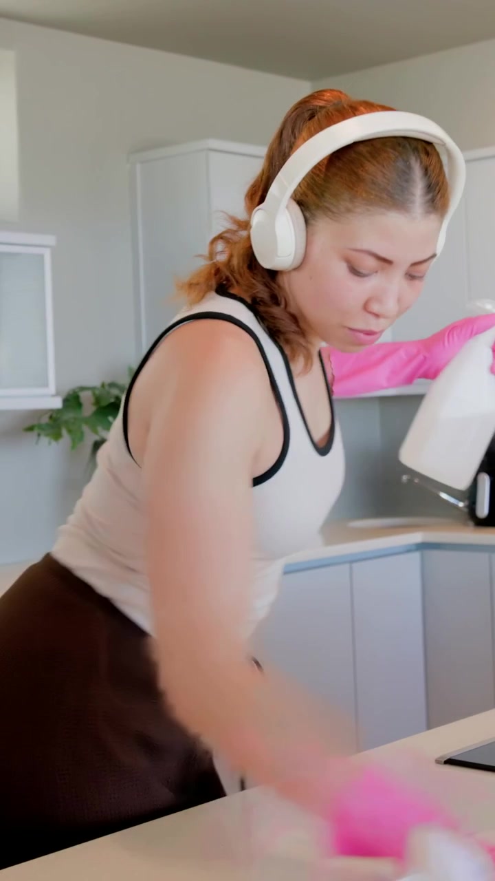 Video verticale: Donna con guanti rosa e cuffie che alza le braccia, spruzza e pulisce il bancone della cucina.