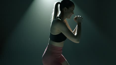 blonde fit woman wearing pink shorts and black sports bra performs side lunges with resistance band. she smiles and approaches the camera. upper body only, dramatic light in smoky gym