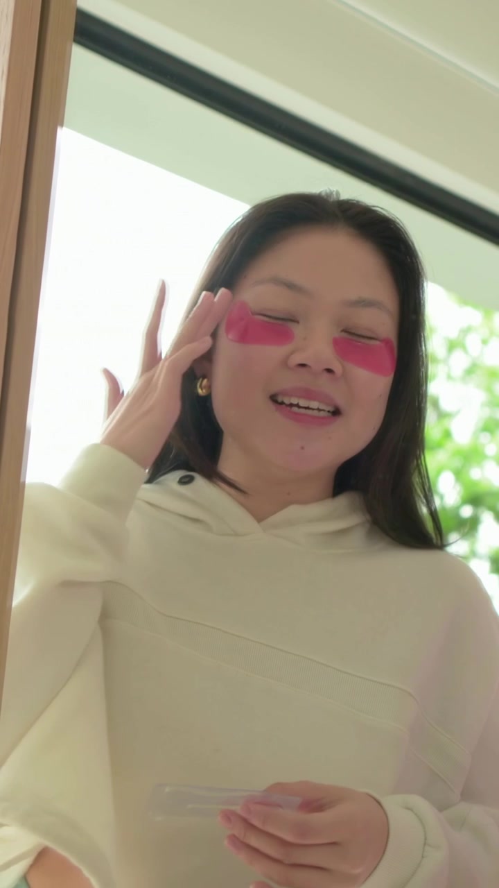Vertical video: Opening packet woman in hoodie peeling and pressing pink eye pads at home for eyes