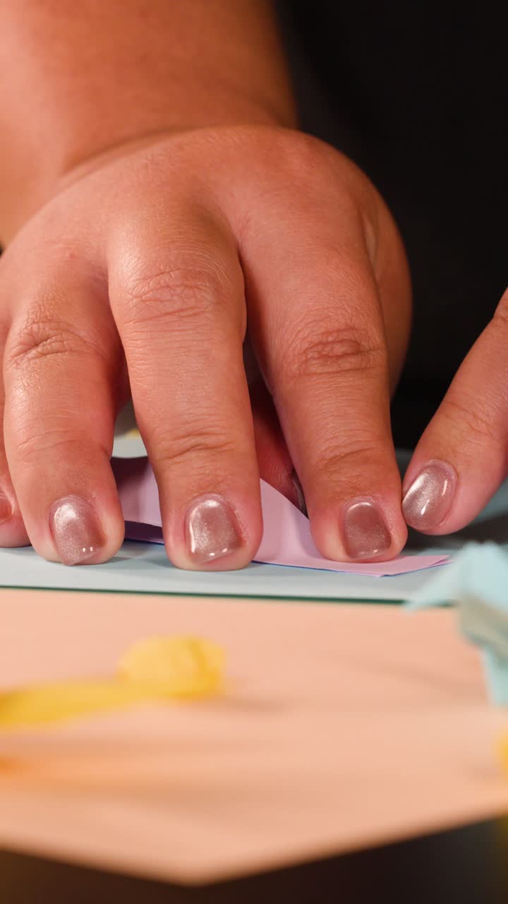 Close-up shot of hands with metallic nail polish precisely folding pink paper on blue surface