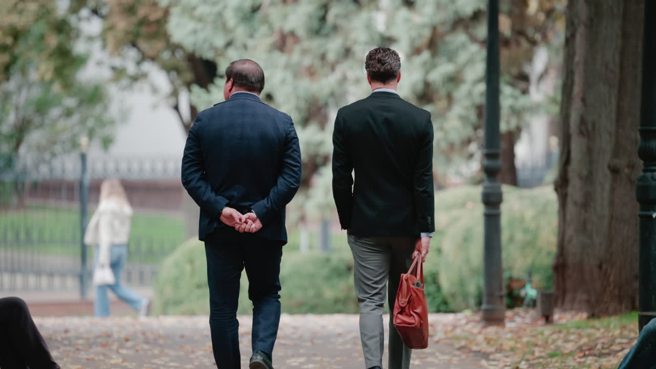 Two men in suits walk together through a leafy park, engaged in a business conversation during a calm autumn day