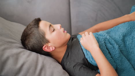 side view of sick boy covered with blue blanket, adjusting on couch while coughing covering his mouth, looking uncomfortable, resting indoors