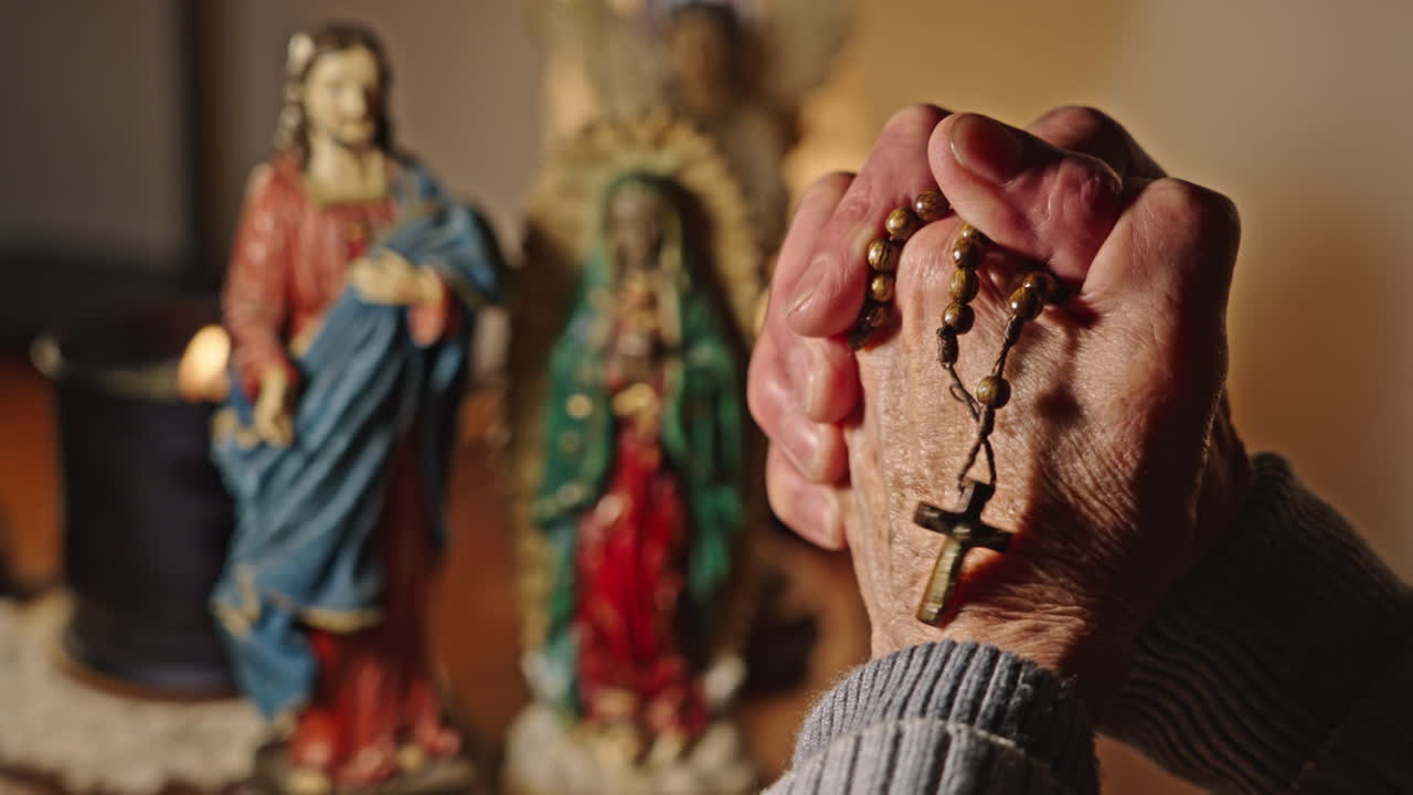Primo piano di mani in preghiera con rosario e statue religiose.