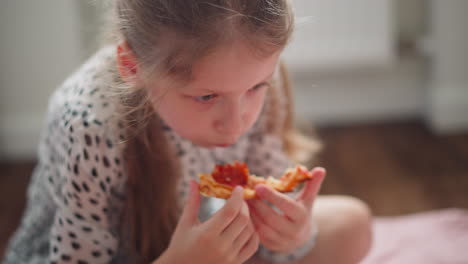 close up little girl holding and eating cheesy pepperoni pizza with both hands, focused expression on face, seated on wooden floor, blurred background