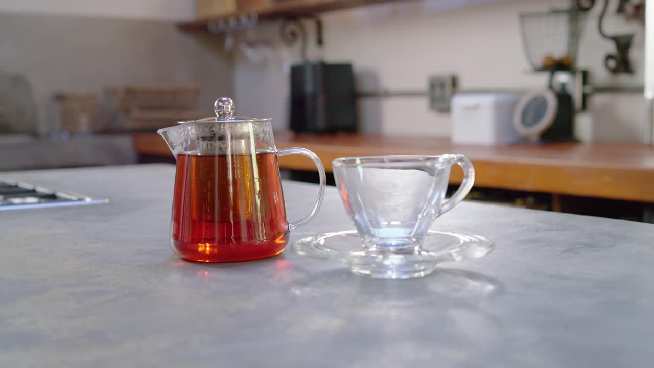 La telecamera si allontana e si inclina verso l'alto, rivelando una teiera di vetro e una tazza da tè vuota su un bancone grigio.