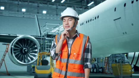 ingeniero masculino asiático con casco de seguridad de pie con aviones en el hangar. pensando en algo y mirando a su alrededor mientras el mantenimiento de aviones