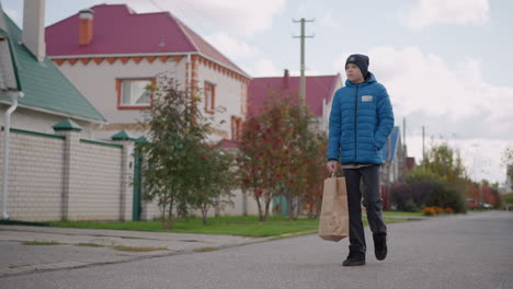 little boy in blue jacket strolling on residential street with thoughtful look, carrying paper bag, blurred background features houses, trees, and greenery