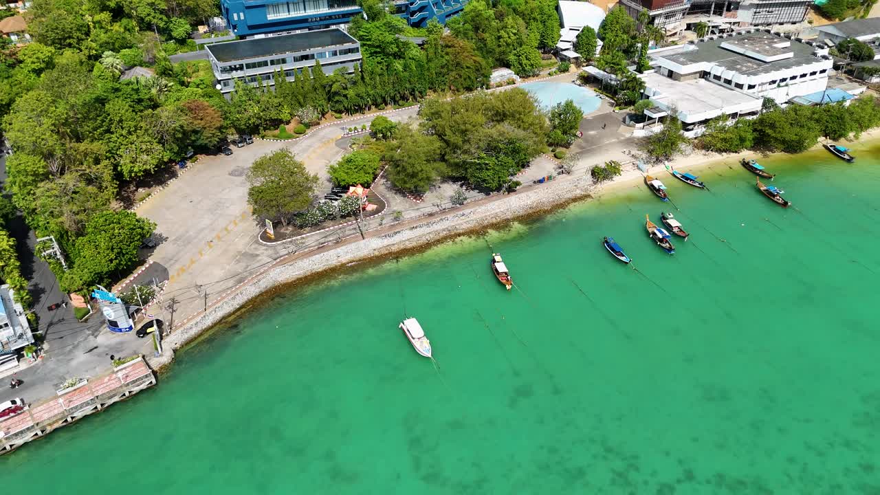 Drone footage captures longtail boats along Phuket's Patong Beach. Vibrant turquoise waters and lush greenery create a serene coastal scene