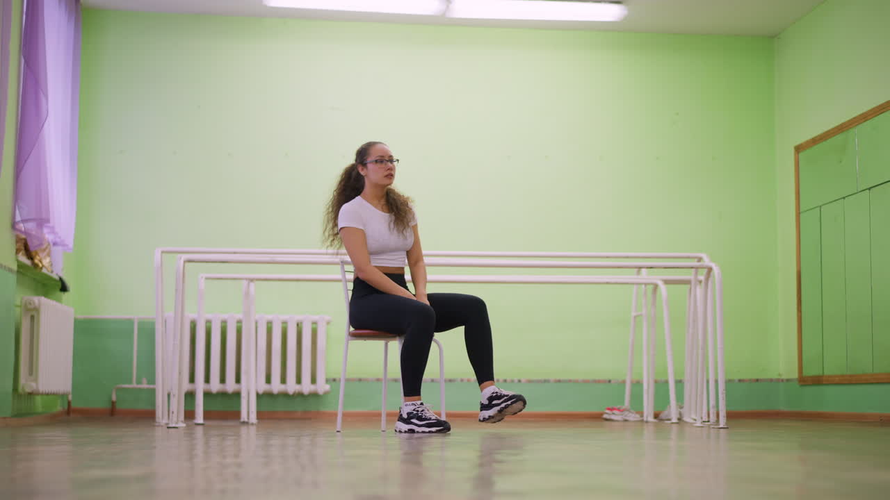 Girl in white top and black leggings with canvas sneakers sitting on chair inside studio, appearing stressed and exhausted after workout, wooden floor and mirror reflection completing indoor