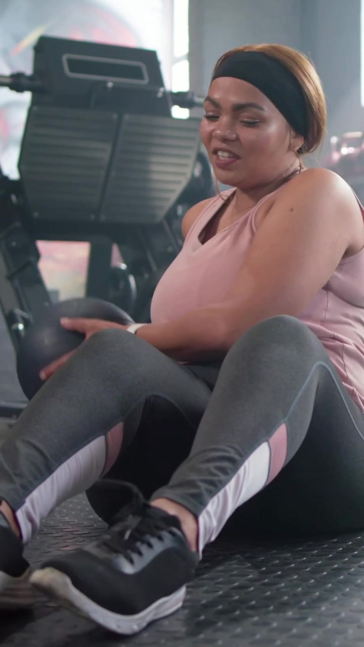 Video verticale: Una donna con un top rosa tiene una palla medica nera e fa torsioni del busto in palestra, con un partner che si unisce.