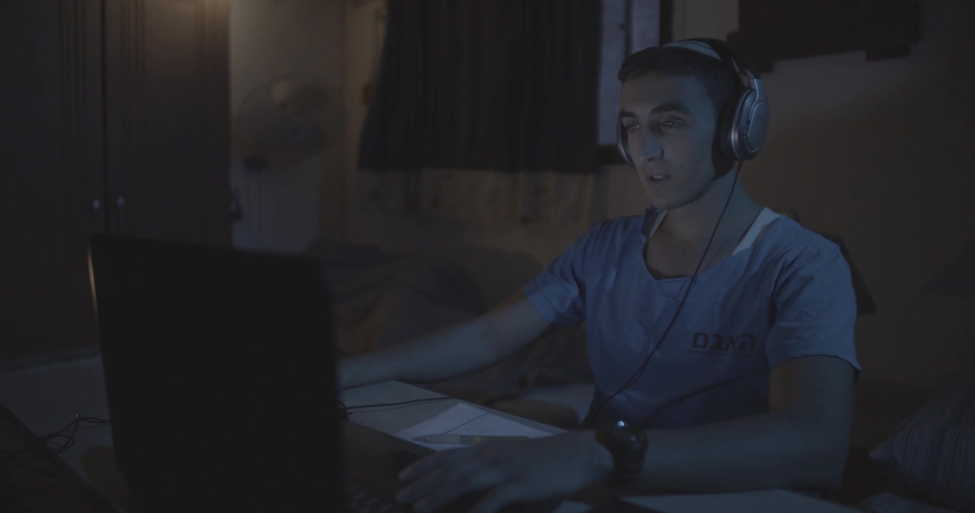 A young man wearing a light blue T-shirt sits in the dark at night in front of a computer with headphones in a room at a table