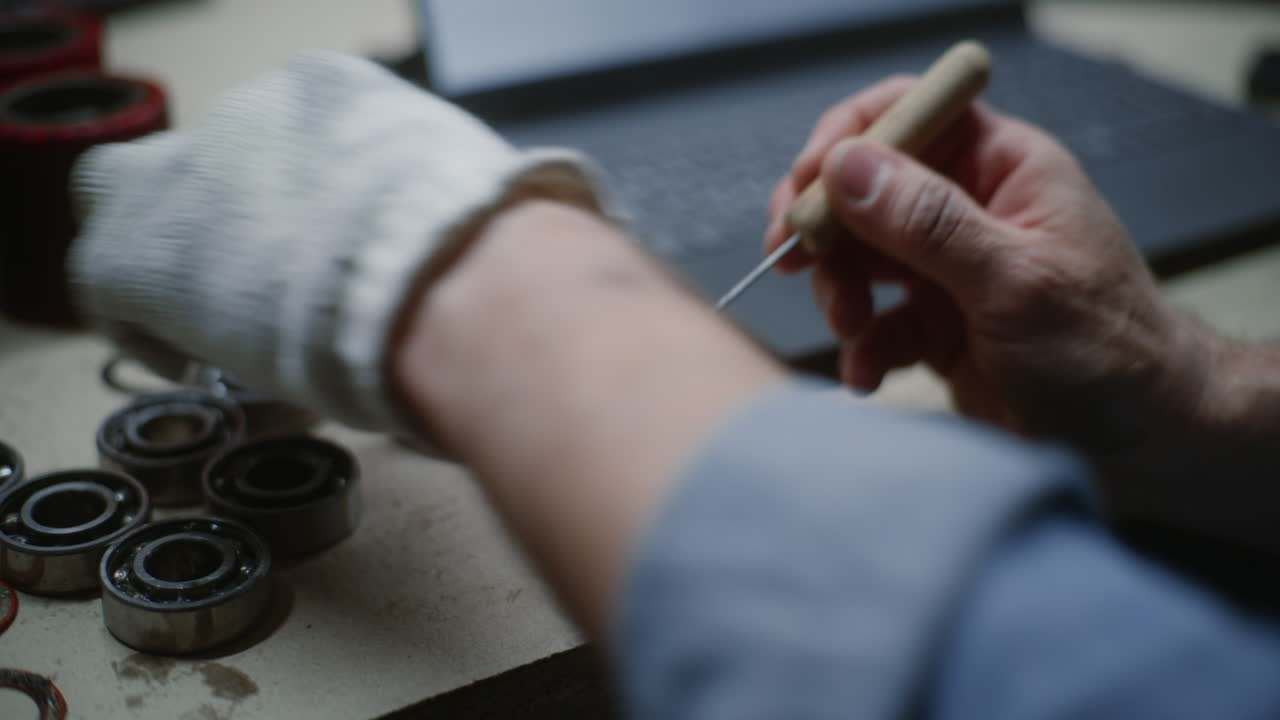 During Working Day in Workshop: Close Up of Craftsman, Maintenance Worker Doing Handwork. Man Cleaning or Servicing Bearings, Combining Work with Online Studying. In the Background Unfocused Laptop.