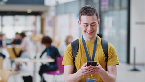 smiling male college student checking mobile phone in busy communal campus building