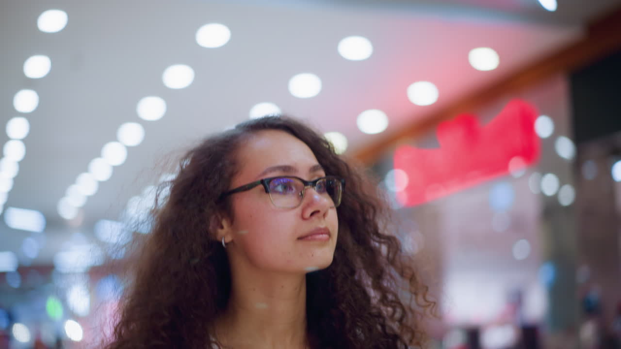 Woman with glasses and curly hair looking around in well-lit mall, illuminated by soft bokeh lights in background and people walking in the mall blurred