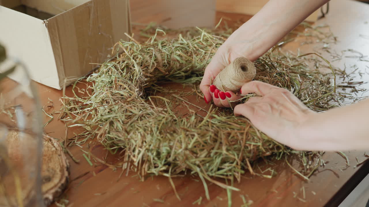 Hands with red nails tying twine around straw wreath on wooden table, crafting rustic decoration using dried grass, cardboard box nearby, creative handmade process showing natural materials