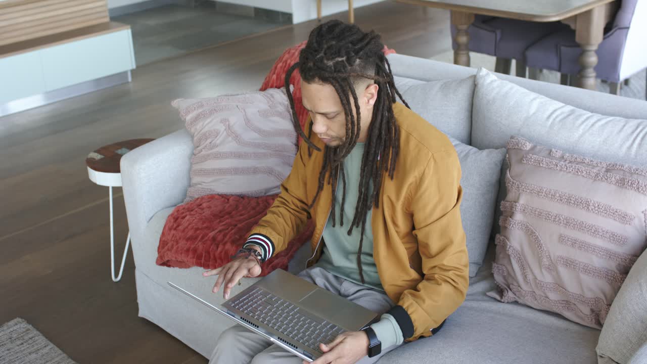 Middle-aged man entering living room, sitting on sofa, placing laptop on lap, opening lid, working