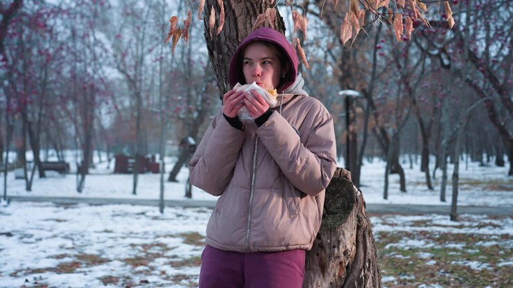 giovane ragazza in abito invernale che si riposa sull'albero che mangia spuntini nel parco all'aperto durante l'inverno, riflesso di luce dallo sfondo, che si gode il momento del cibo nel parco, le vibrazioni del tempo freddo