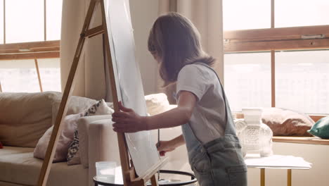 side view of a blonde girl painting her drawing with a brush on a lectern in the living room at home