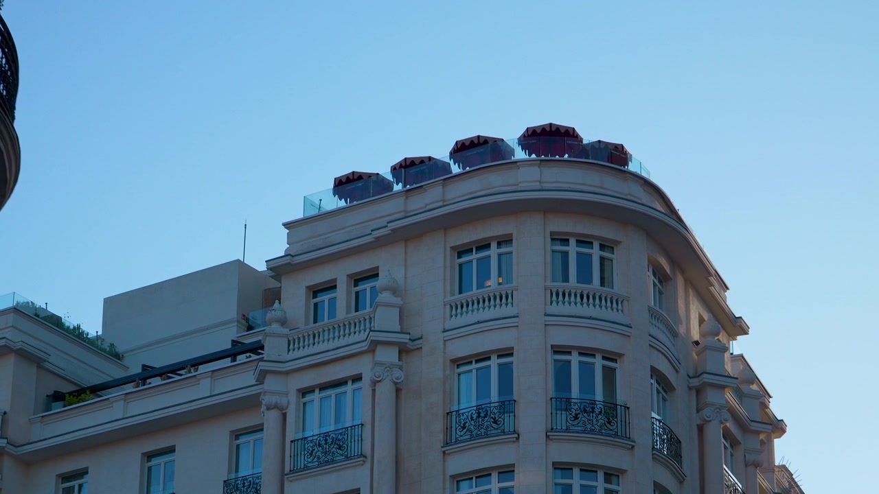 Vista dal basso della facciata di un edificio storico europeo a Madrid, caratterizzata da balconi ornati e una moderna terrazza panoramica con ombrelloni rossi contro un cielo azzurro limpido