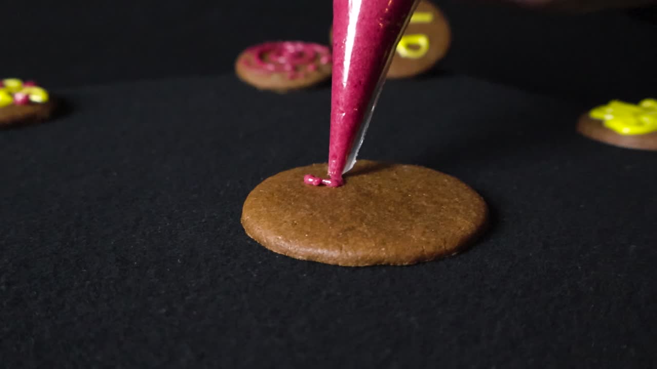 Gingerbreads being decorated in slow motion with purple or red vibrant sugar coating coming out of a nozzle piping tube. The cookies are placed on a dark black studio background under good lights.