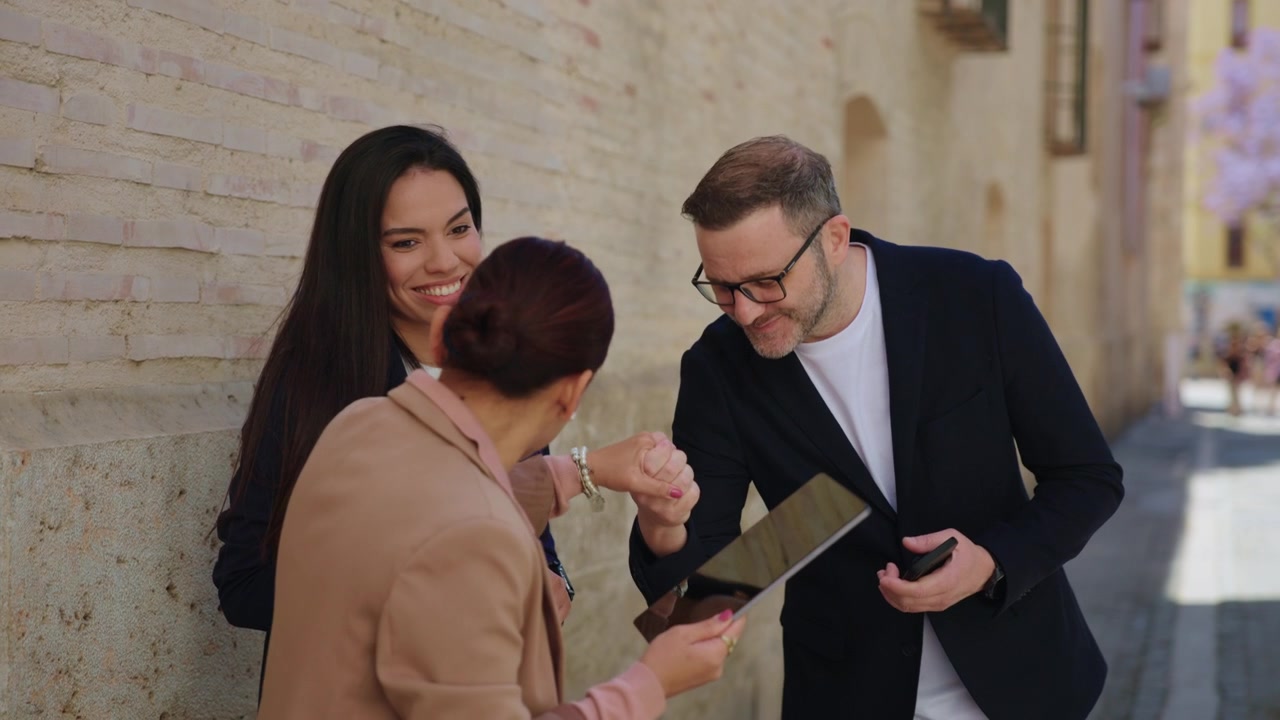 Um empresário se aproxima de duas colegas que revisam um tablet digital ao ar livre, apertando as mãos e cumprimentando-as respeitosamente em um cenário de cidade histórica.