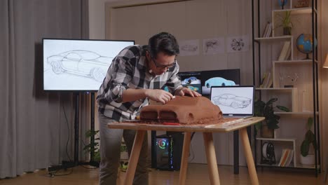 hombre asiático diseñador de automóviles con una computadora portátil usando rastrillo o alambre para suavizar la superficie y crear detalles en la escultura de arcilla de automóvil en el estudio