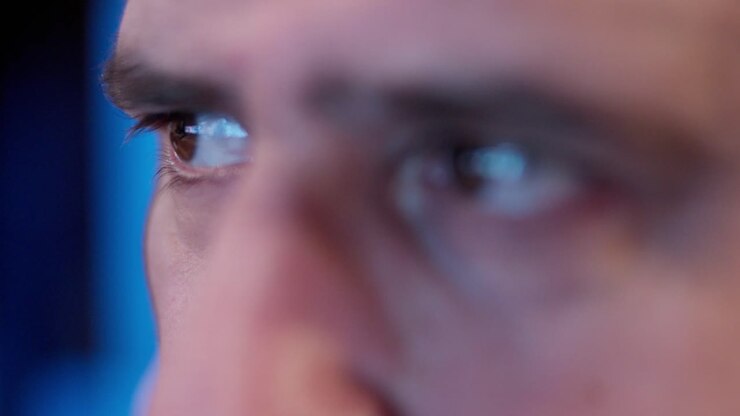 Close view of a male face showing intense eyes looking to the side. Blue lighting reflects on the skin, indicating a screen or monitor environment during work or focus. Cinematic close-up