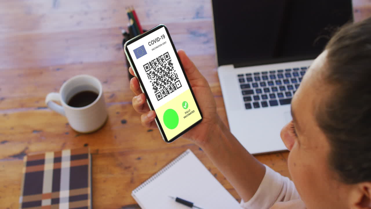 Woman at desk holding smartphone with covid vaccination certificate, eu flag and qr code on screen