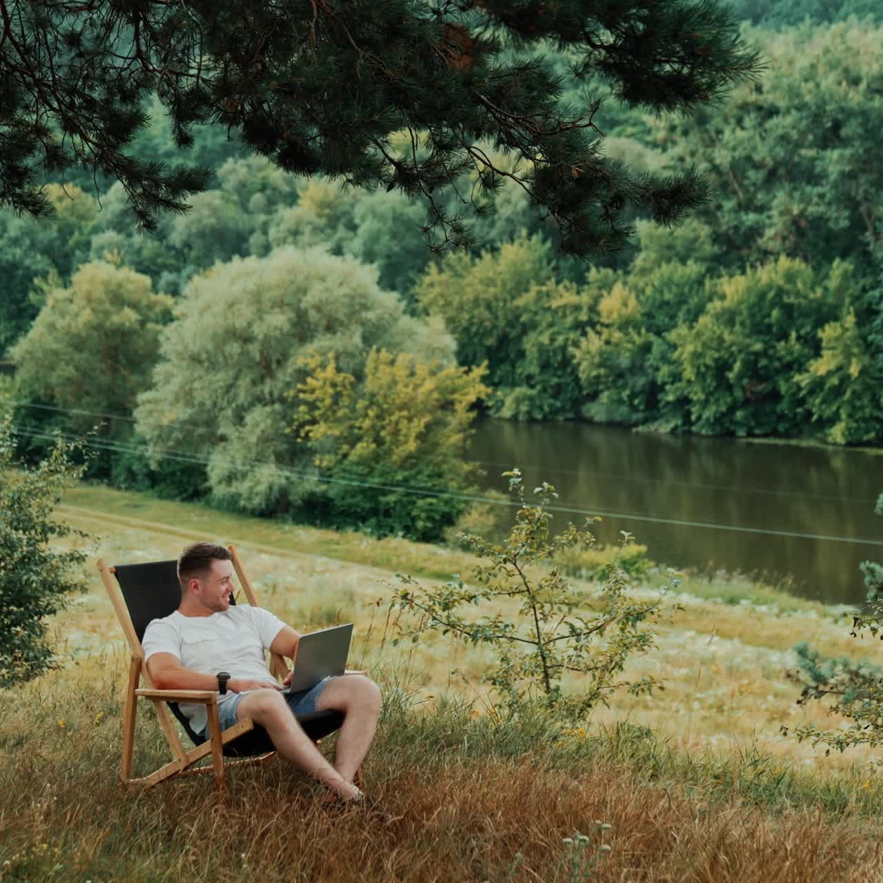 Remote work in the nature away from the office. Caucasian smiling man is in folding chair holding his laptop